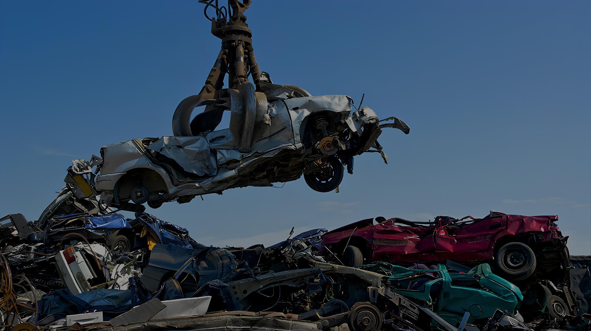 Schrottplatzgreifer mit 4 Zinken hebt gepresstes Auto in die Luft. Am Boden liegen weitere gepresste Autos